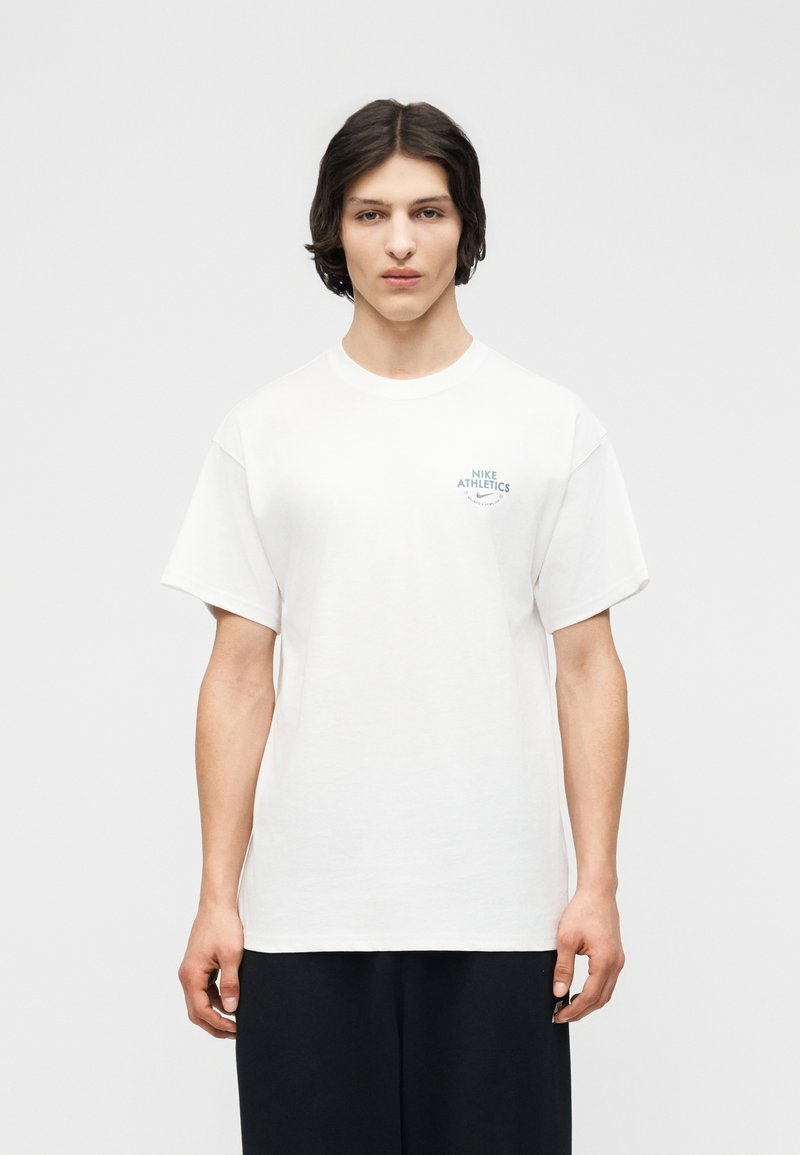 Giovane adulto con capelli castani di media lunghezza che indossa una maglietta bianca Nike Athletics e pantaloni neri, in piedi davanti a uno sfondo semplice.