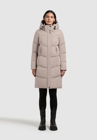 Gaiši bēšs pūķa mētelis ar augstu apkakli, horizontālu šūtu rakstu un divām sānu kabatām. Kombinēts ar melnām legingām un masīvām melnām zābakiem.