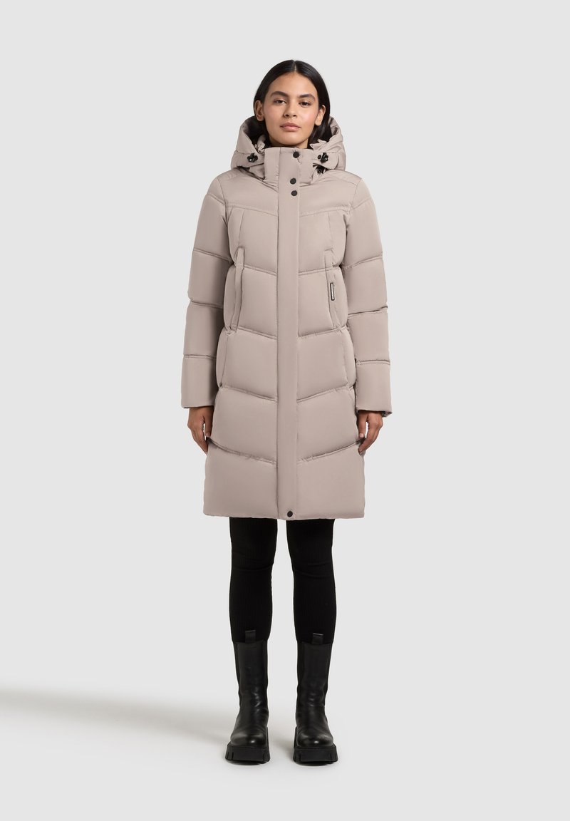 Gaiši bēšs pūķa mētelis ar augstu apkakli, horizontālu šūtu rakstu un divām sānu kabatām. Kombinēts ar melnām legingām un masīvām melnām zābakiem.