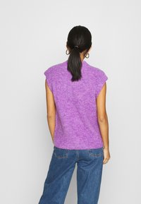 Purple knitted sleeveless top with a textured finish, wide shoulder seams, and a relaxed fit, paired with high-waisted blue jeans.