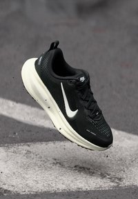 Mustat juoksukengät, joissa on hengittävä verkkomateriaali, valkoinen Nike-tikkaus, teksturoitu yksityiskohta ja pehmustettu pohja, suspendoituna pinnan ylle.