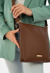 Bruine leren crossbodytas met een getextureerd oppervlak, met zijstiksels, gouden hardware en een rechthoekige vorm, vastgehouden door een persoon.