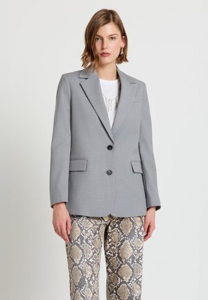 Frau trägt ein hellgraues Blazer über einem weißen Shirt mit einer silbernen Halskette und beige-grau gemusterte Schlangenprint-Hose vor einem einfarbigen Hintergrund.