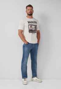 Crème kortemou t-shirt met een grafisch ontwerp van een oude televisie met tekst; gecombineerd met lichtblauwe denim jeans en beige sneakers.