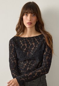 Czarna koronkowa bluzka z długim rękawem z kwiatowym wzorem, z halsztuką, oraz teksturowanymi rękawami, w połączeniu z dopasowanym szarym pasem.