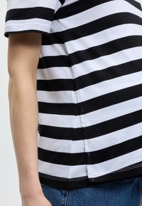 Χέρι και κορμός ατόμου με κοντομάνικη μπλούζα σε μαύρες και άσπρες οριζόντιες ρίγες και μπλε τζιν, μπροστά σε απλό φόντο.