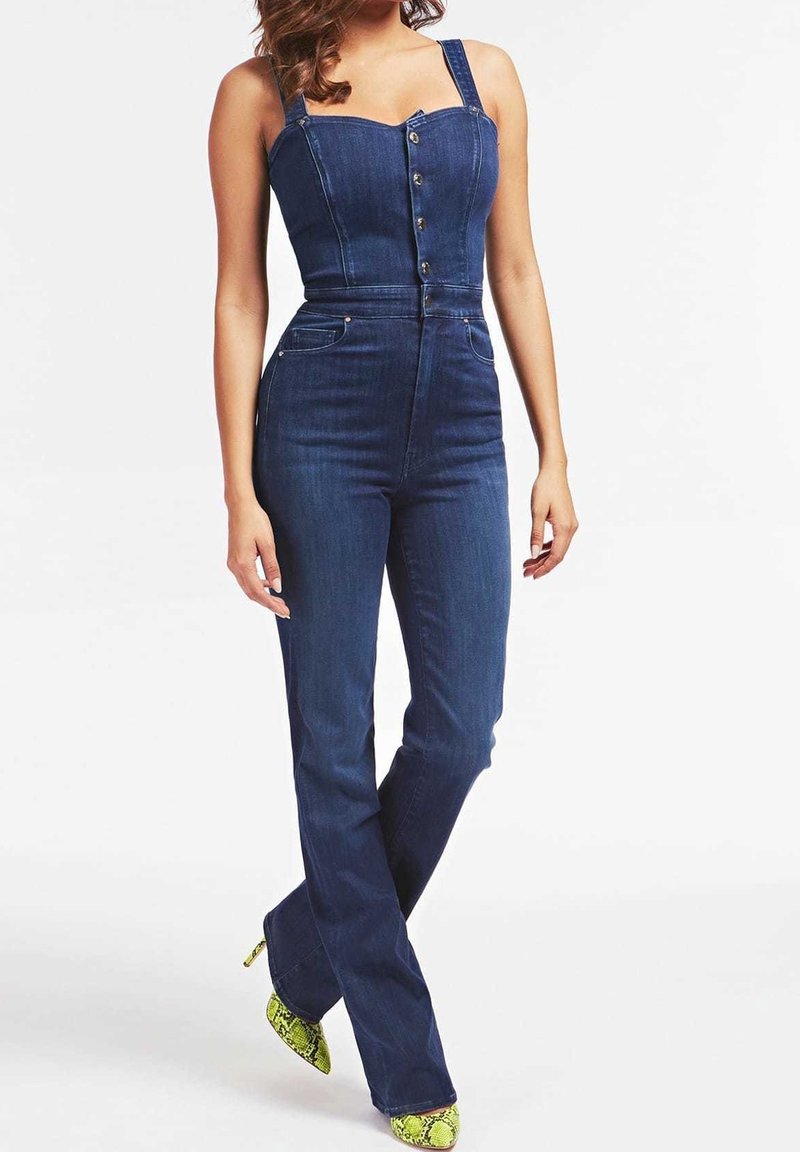 Femme portant une combinaison en denim bleu foncé ajustée avec des boutons sur le devant et des chaussures à talons hauts jaunes à imprimé serpent, debout sur fond blanc.