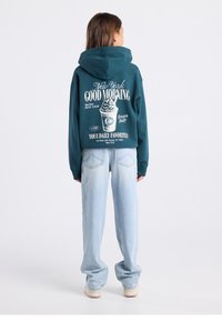 Teal hoodie with large white graphic on back, featuring text and ice cream design. Paired with light blue, relaxed-fit jeans and sneakers.
