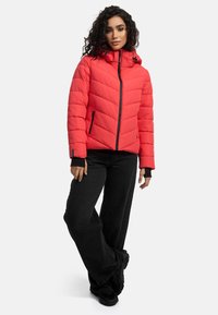 Red puffer jacket with a hood, diamond quilt pattern, zip closure, and black accents. Worn with black wide-leg pants and sneakers.