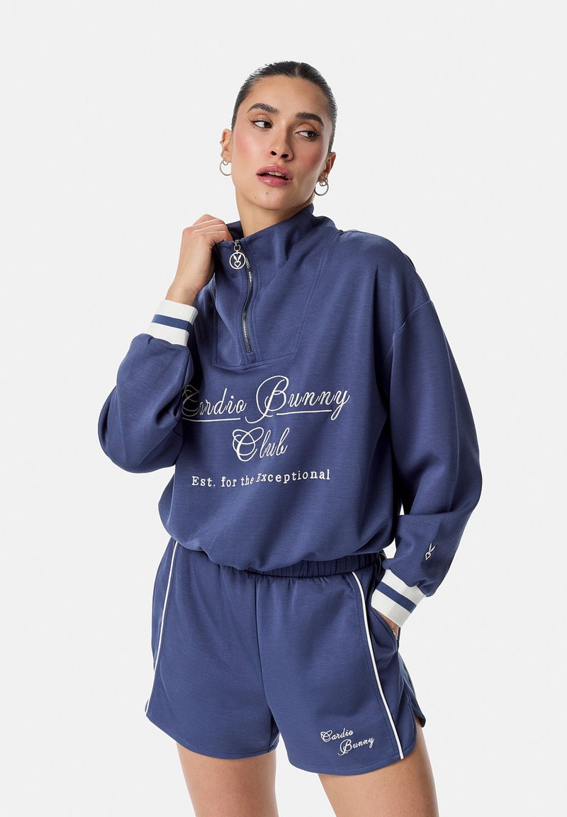 Mujer con conjunto a juego de sudadera y pantalones cortos azul marino con texto blanco y puños a rayas, posando con la mano cerca del cuello.