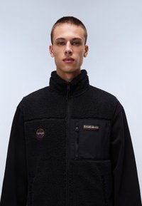 Black fleece vest with a high collar, zippered chest pocket, and textured fabric on the front. Features a logo patch on the upper left.