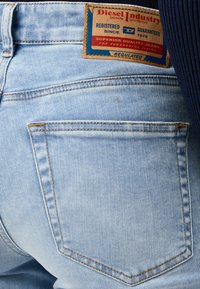 Ljusblå denimjeans med texturerad yta, featuring en brun läderlogga och två bakfickor med gula sömnadsdetaljer.