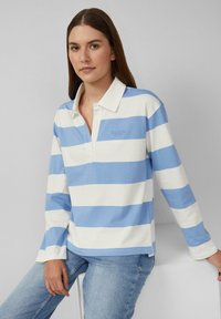 Frau in einem blau-weißen, horizontal gestreiften Langarm-Polohemd und hellblauen Jeans, die auf einem weißen Block vor grauem Hintergrund sitzt.