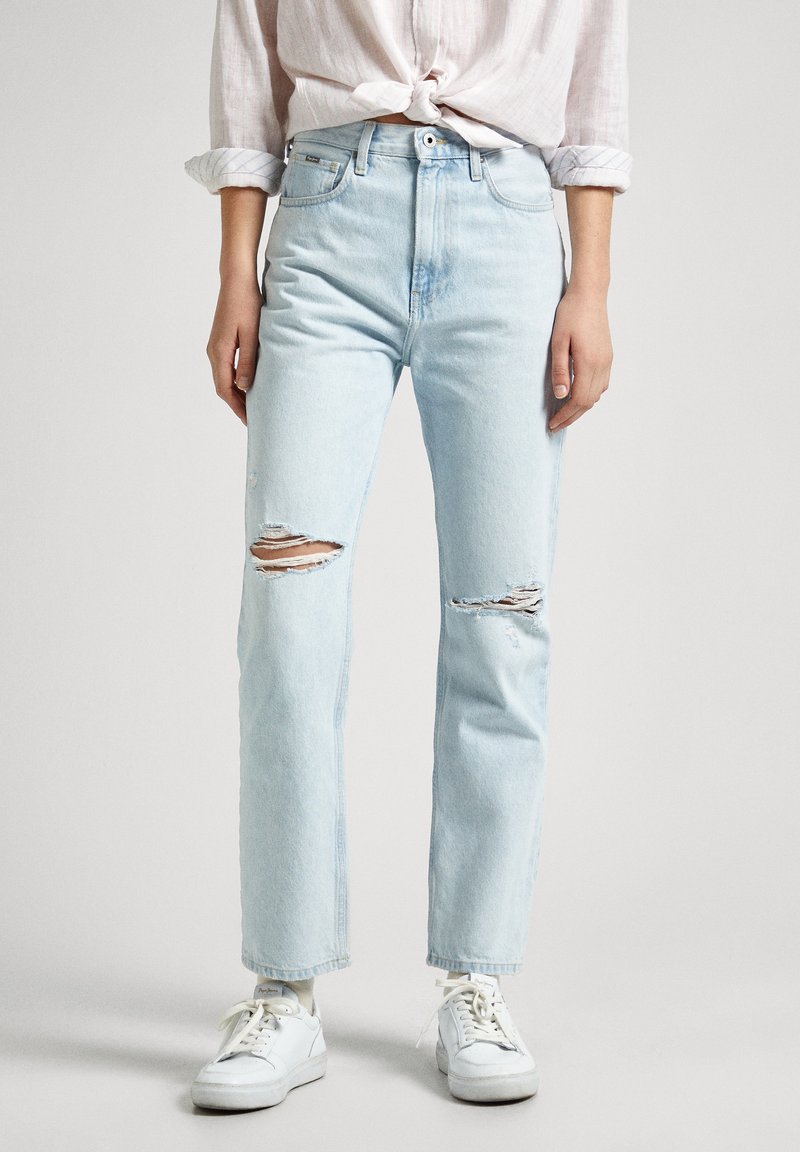 Persona que lleva puestos unos jeans claros desgastados, zapatillas blancas y una camisa de un color claro con las mangas enrolladas atadas a la cintura, de pie contra un fondo liso.