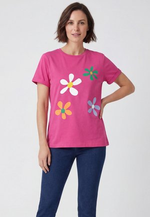 Donna che indossa una maglietta rosa con grandi stampe floreali colorate, abbinata a jeans blu, in piedi con una mano sui fianchi.