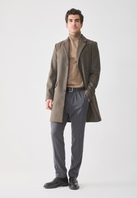 Hombre de pie con un abrigo marrón, suéter de cuello alto color beige, pantalones grises, cinturón negro y zapatos de vestir negros frente a un fondo liso.