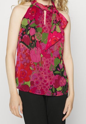 Kvinde iført ærmeløs halterneck-top med pink, rød, lilla og grøn blomsterprint og bindebånd ved halsen, kombineret med sorte bukser.