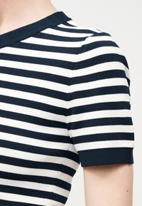 Gros plan sur l'épaule d'une personne portant un t-shirt en maille à manches courtes rayé horizontalement noir et blanc avec un col rond.