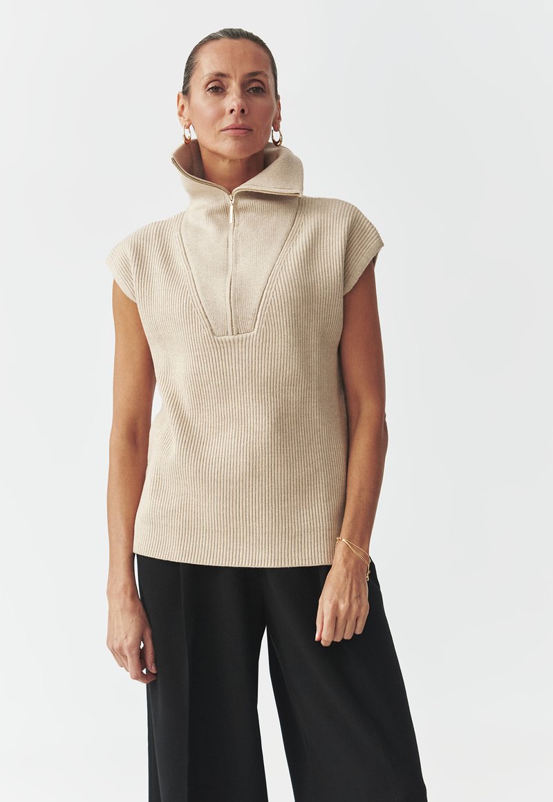 Beige ribbad ärmlös topp med hög krage och dragkedjestängning, med en djup v-ringning och textilstruktur. Matchad med svarta vida byxor.