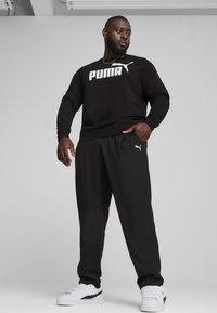 Čierna mikina s dlhým rukávom a bielym logom "PUMA", skombinovaná s voľnými čiernymi nohavicami a bielymi teniskami s čiernymi akcentmi.