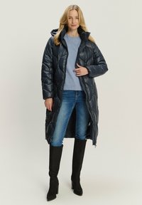 Marineblauer, gesteppter Puffer-Mantel mit Kapuze, langes Design und Reißverschluss, kombiniert mit einem leichten Pullover und blauen Jeans, getragen mit kniehohen schwarzen Stiefeln.