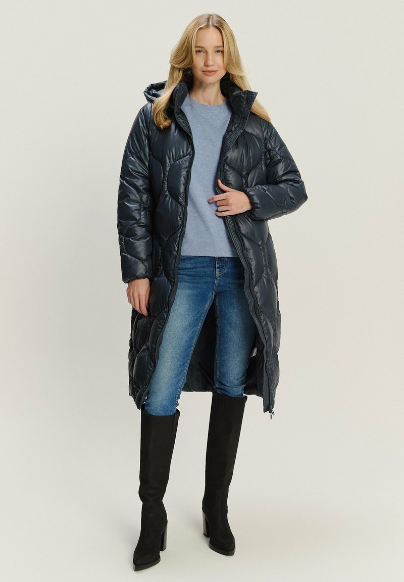 Marineblauer, gesteppter Puffer-Mantel mit Kapuze, langes Design und Reißverschluss, kombiniert mit einem leichten Pullover und blauen Jeans, getragen mit kniehohen schwarzen Stiefeln.