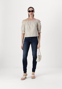 Off-shoulder, beige mönstrad topp med volangkrage, ihop med mörkblå skinnyjeans och vita klacksandaler, hållande en liten väska.