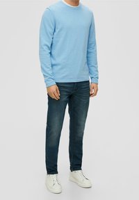 Man wearing light blue long-sleeve shirt, dark blue jeans, and white sneakers standing against a plain background.