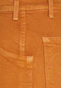 Oranger Denimstoff mit einer Vordertasche, sichtbaren Nähten und einer strukturierten Oberfläche. Eine zweite kleinere Tasche befindet sich oberhalb der Haupttasche.