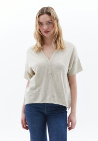 Helles beige kurzärmeliges V-Ausschnitt-Oberteil mit fünf Knöpfen und rohem Saum. Weicher Stoff, lockere Passform, kombiniert mit blauen Jeans.