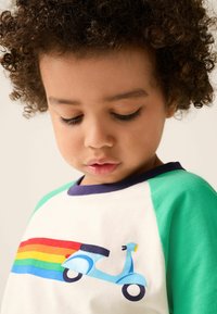 Balta T-krekls ar zaļām piedurknēm, uz kura attēlots zils skūteris un varavīksnes svītras. Īsas piedurknes, apaļa kakla izgriezuma forma un gluda kokvilnas tekstūra.