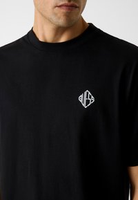 Schwarzes Baumwoll-T-Shirt mit rundem Ausschnitt und weiß gesticktem "GUESS"-Logo in Rautenform auf der linken Brust. Kurze Ärmel.