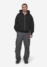 Črna jopa s kapuco z zadrgo in sprednjimi žepi ter sive cargo hlače z več žepi in nastavljivimi robovi. Nošeno z temnimi supergami.