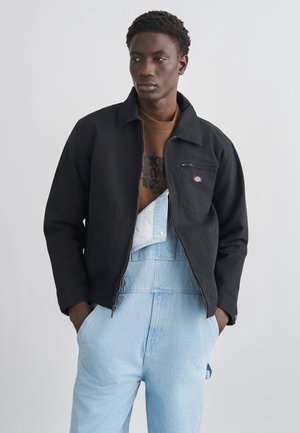 Młody mężczyzna ubrany w jasnoniebieskie ogrodniczki jeansowe, brązową bluzę z nadrukiem oraz czarną roboczą kurtkę zapinaną na suwak, stojący z rękami w kieszeniach.