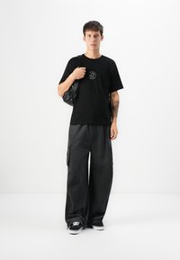 Schwarzes Baumwoll-T-Shirt mit einem runden Grafikdesign, kombiniert mit weit geschnittenen, dunkelgrauen Cargo-Hosen mit Taschen und schwarzen Sneakern mit weißen Sohlen.