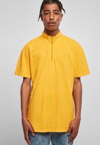 Gul kortermet topp med høy krage og halv glidelås, som har en glatt tekstur og en avslappet passform. Kombinert med lys blå jeans.