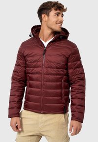 Jachetă puffer burgundy cu fermoar frontal, design matlasat și glugă. Dispune de două buzunare laterale și un logo pe mânecă.