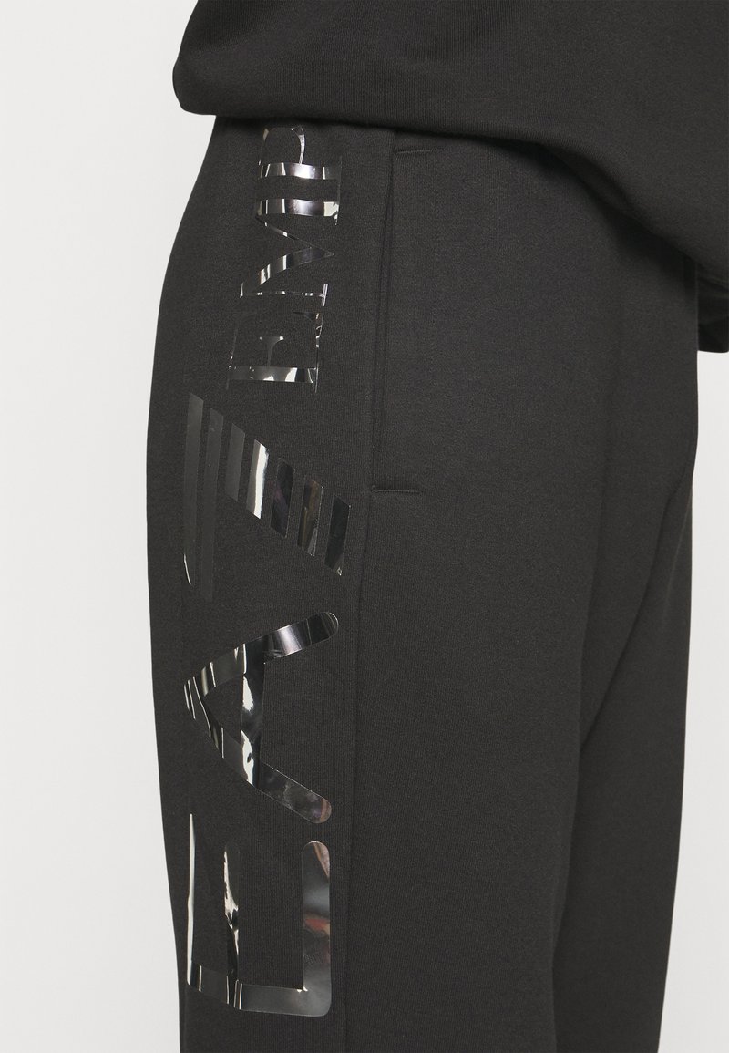 Pantalones de chándal negros con acentos metálicos reflectantes en los laterales, que cuentan con elementos gráficos audaces y un ajuste estructurado y cómodo.