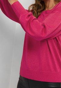 Pull en maille fuchsia à manches longues, avec poignets côtelés et coupe décontractée. Le tissu présente une légère brillance, ajoutant de la texture au design.