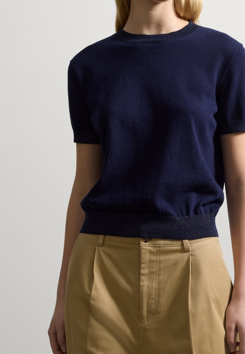Femme portant un haut en maille à manches courtes bleu marine ajusté, rentré dans un pantalon beige à taille haute, sur un fond neutre.