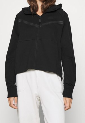 Personne portant une veste à capuche noire zippée avec des manches longues et un pantalon blanc, vue du cou jusqu'aux hanches sur un fond uni.