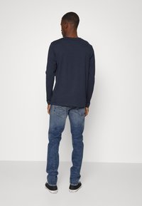 Camisa de manga larga azul marino, mezcla de algodón, con cuello redondo. Combinada con jeans azules y zapatillas negras. Costuras visibles y bolsillos de los jeans.