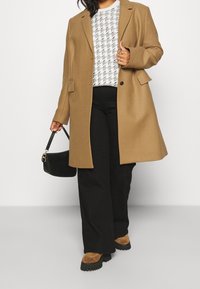 Personne portant un manteau camel, un pantalon noir à jambes larges, un pull blanc à motif, des chaussures brunes à semelle épaisse, et tenant un sac à main noir.