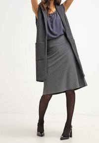 Gilet senza maniche grigio con tasche frontali, abbinato a una gonna grigia fino al ginocchio. Tacchi a spillo neri lucidi e calze scure completano l'outfit.