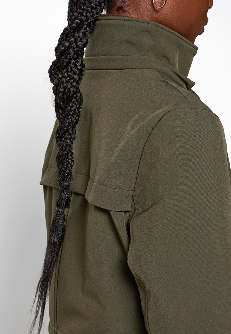 Giacca verde oliva con colletto alto, dotata di tasche a soffietto, una texture liscia e un design aderente. Capelli lunghi e intrecciati visibili nell'inquadratura.