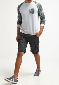 Uomo che indossa una felpa grigia con maniche e tasca verde scuro a palme, pantaloni cargo neri e sneakers grige mentre cammina in avanti.