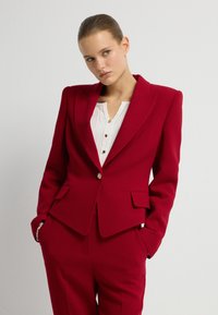 Žena s hladce Back vlasy, oblečená v červeném střiženém blejzru a odpovídajících kalhotách, s rukama v kapsách, mírně se dívá stranou na neutrálním pozadí.