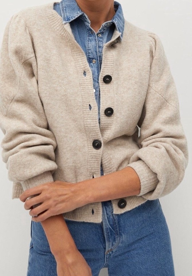 Cardigan en laine beige avec un col rond, cinq boutons noirs et des manches bouffantes, superposé à une chemise en denim bleu.