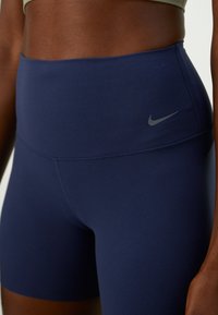 Pogled izbliza na osebo, oblečeno v športne kratke hlače za visoko pasom v mornarsko modri barvi z majhnim temnosivim Nike logotipom na levem bok. 