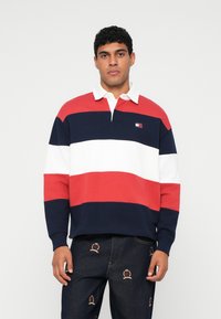 Poloshirt met lange mouwen in rood, wit en marineblauw met horizontale strepen. Heeft een witte kraag en een klein logo op de borst. Draagt bij broeken met borduursel.